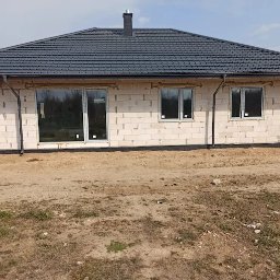 F.H.U Malors Marcin Słomski - Surowy dom z białych bloczków z ciemnym dachem i oknami, widoczny z zewnątrz na tle nieba. Wokół domu piasek i trawa, widoczny komin.