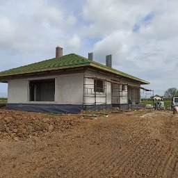 F.H.U Malors Marcin Słomski - Niedokończony dom jednorodzinny z zielonym dachem i widocznymi rusztowaniami. W tle krajobraz z polami i zabudowaniami. Prace budowlane w toku.