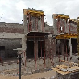 F.H.U Malors Marcin Słomski - Budowa domu w trakcie: murowane ściany z otworami okiennymi i elementami szalunków. Na górze budynku widoczna osoba w kurtce. Na pierwszym planie składowane płyty.