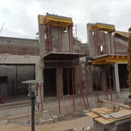 Budowa domu w trakcie: murowane ściany z otworami okiennymi i elementami szalunków. Na górze budynku widoczna osoba w kurtce. Na pierwszym planie składowane płyty.