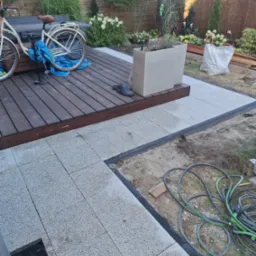 Nowa, jasnoszara kostka brukowa na podwórku, z widocznym odpływem liniowym i fragmentem drewnianego tarasu z rowerem. W tle roślinność ogrodowa.