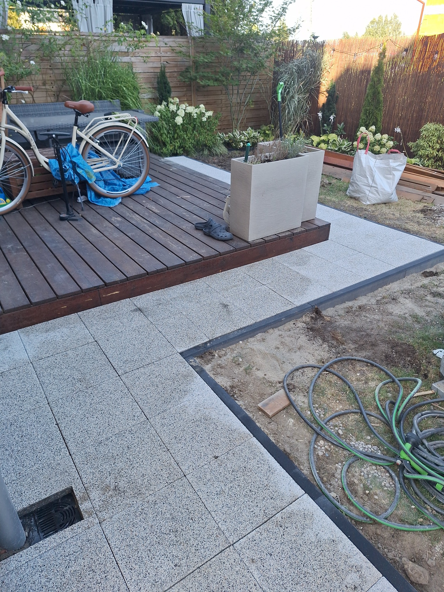 Nowa, jasnoszara kostka brukowa na podwórku, z widocznym odpływem liniowym i fragmentem drewnianego tarasu z rowerem. W tle roślinność ogrodowa.