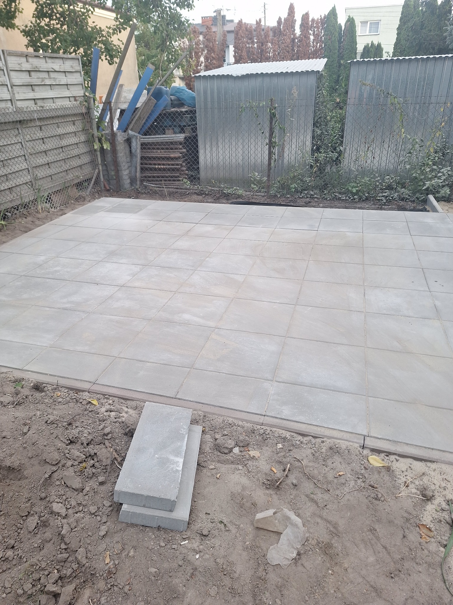 Wyłożony plac z jasnoszarej kostki brukowej na posesji, widoczne niezakończone prace i przygotowane płyty. W tle ogrodzenie z siatki i blaszane zabudowania.