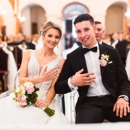 Fotografia Dawid Olkowski - Uśmiechnięta para młoda siedzi w kościele, prezentując obrączki. Panna młoda w sukni z bukietem, pan młody w czarnym garniturze z muchą. W tle ławki i goście.