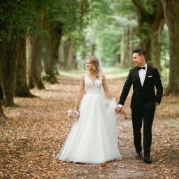 Fotografia Dawid Olkowski - Para młoda spaceruje aleją drzew w jesiennej scenerii. Panna młoda w białej sukni trzyma bukiet, pan młody w czarnym garniturze. Romantyczna atmosfera.