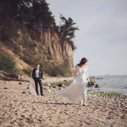 Fotografia Dawid Olkowski - Panna młoda w zwiewnej sukni spaceruje brzegiem morza, trzymając bukiet. W tle klif i elegancki pan młody w smokingu. Romantyczna sceneria ślubna.