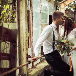Fotografia Dawid Olkowski - Pocałunek pary młodej w zrujnowanej szklarni. Panna młoda w wianku i koronkowej sukni trzyma bukiet. Pan młody w szelkach opiera się o zardzewiałą konstrukcję.