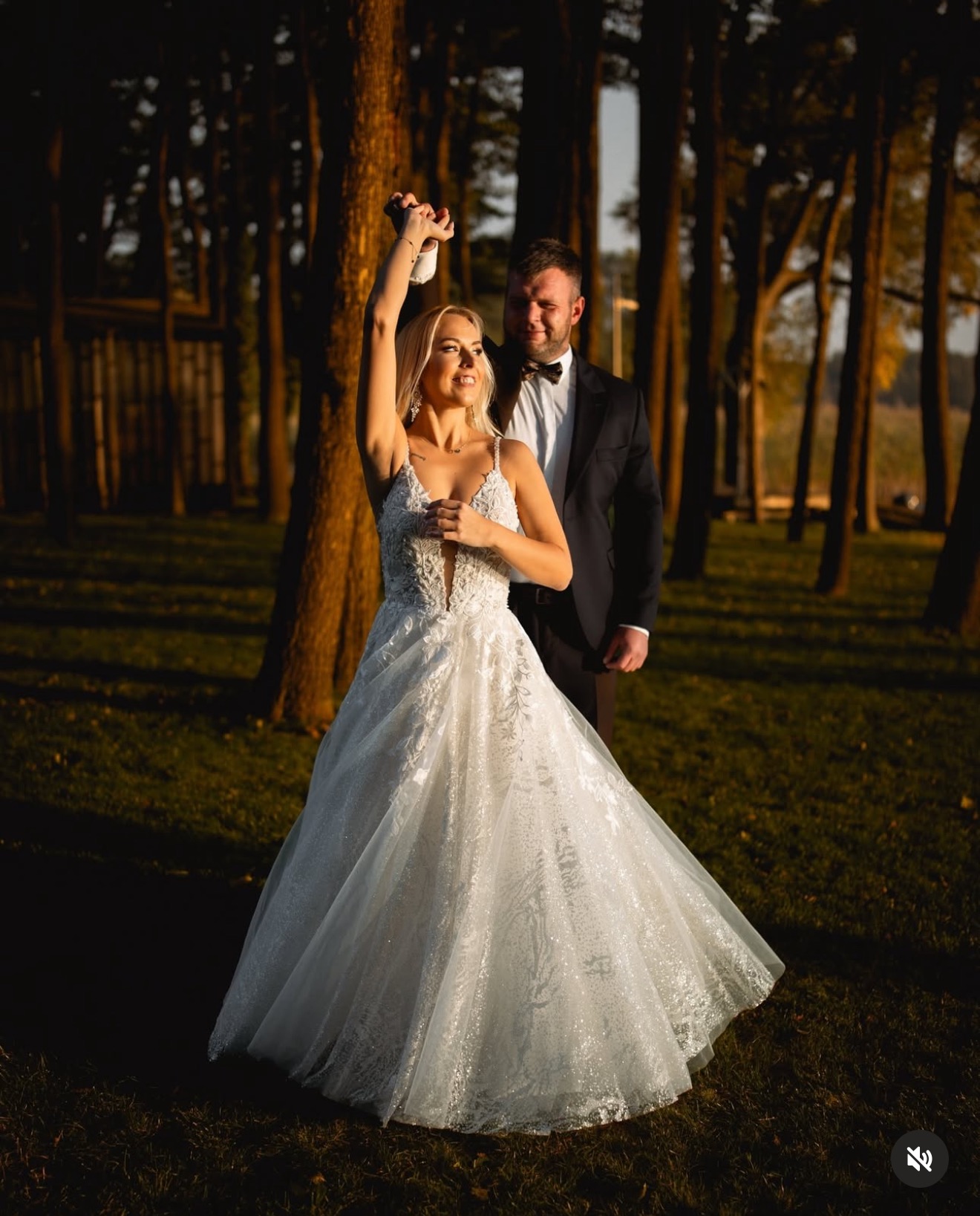 Panna młoda w koronkowej sukni wznosi toast w lesie, obok stoi pan młody w smokingu. Ujęcie w ciepłym, naturalnym świetle.