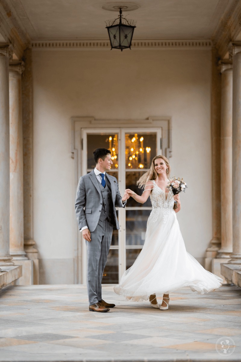 Para młoda trzymająca się za ręce na tle kolumnady i okna z żyrandolem. Panna młoda w koronkowej sukni, pan młody w szarym garniturze. Uśmiechnięci, w ruchu.