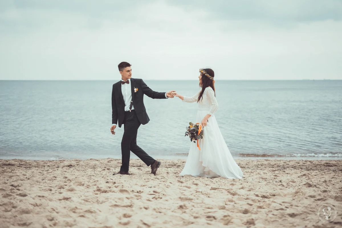Para młoda, on w czarnym garniturze z muchą, ona w białej sukni i wianku, spacerują po piaszczystej plaży, trzymając się za ręce na tle morza i pochmurnego nieba.