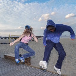 Piotr Domański - Dwie skaczące osoby na tle plaży i mola w słoneczny dzień. Dziewczyna w różowym topie i chłopak w bluzie z kapturem w dynamicznych pozach. W tle widoczni ludzie i elementy plażowej infrastruktury.