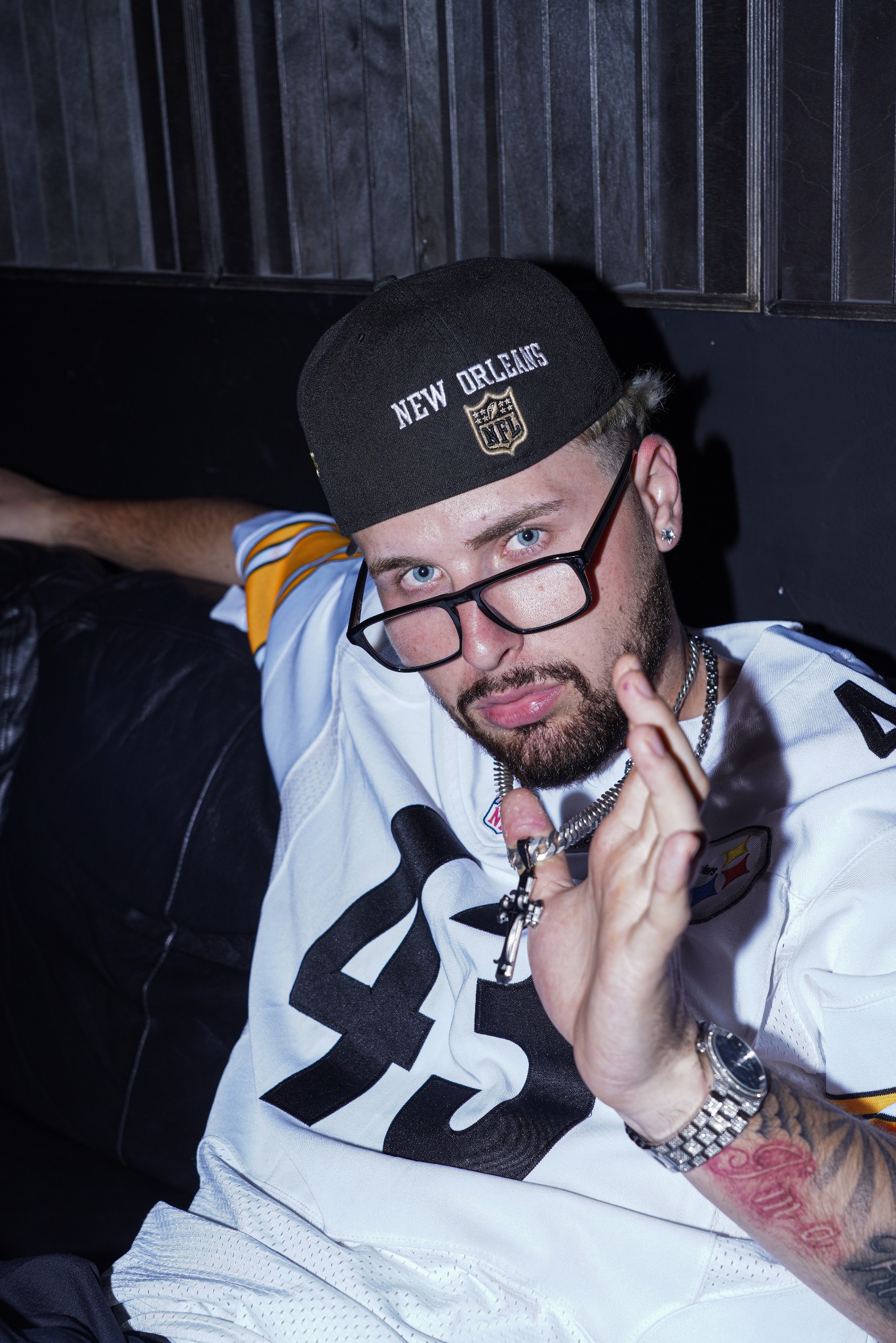 Portret mężczyzny w czapce New Orleans i okularach, z tatuażem na ramieniu, pozującego na tle ciemnej ściany. Ujęcie z bliska, podkreślające detale.