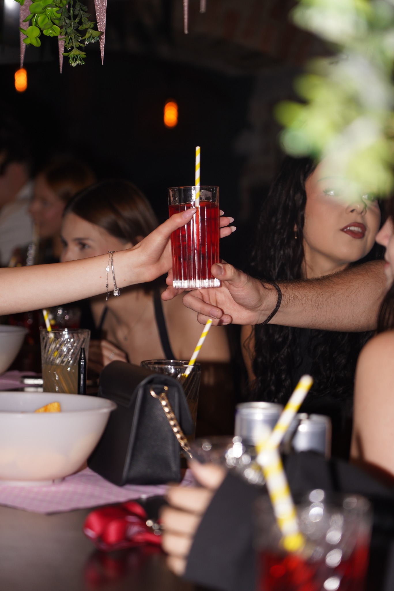 Uroczystość w ciemnych barwach: dłonie podają drinki w szklankach z żółtymi słomkami. W tle uśmiechnięte kobiety i dekoracje.