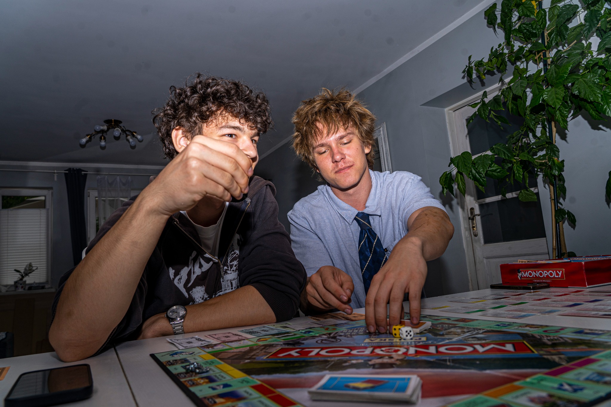 Dwóch młodych mężczyzn gra w Monopoly przy stole w pokoju. Jeden rzuca kostką, drugi trzyma pionek. Widoczna plansza, karty i pudełko gry.