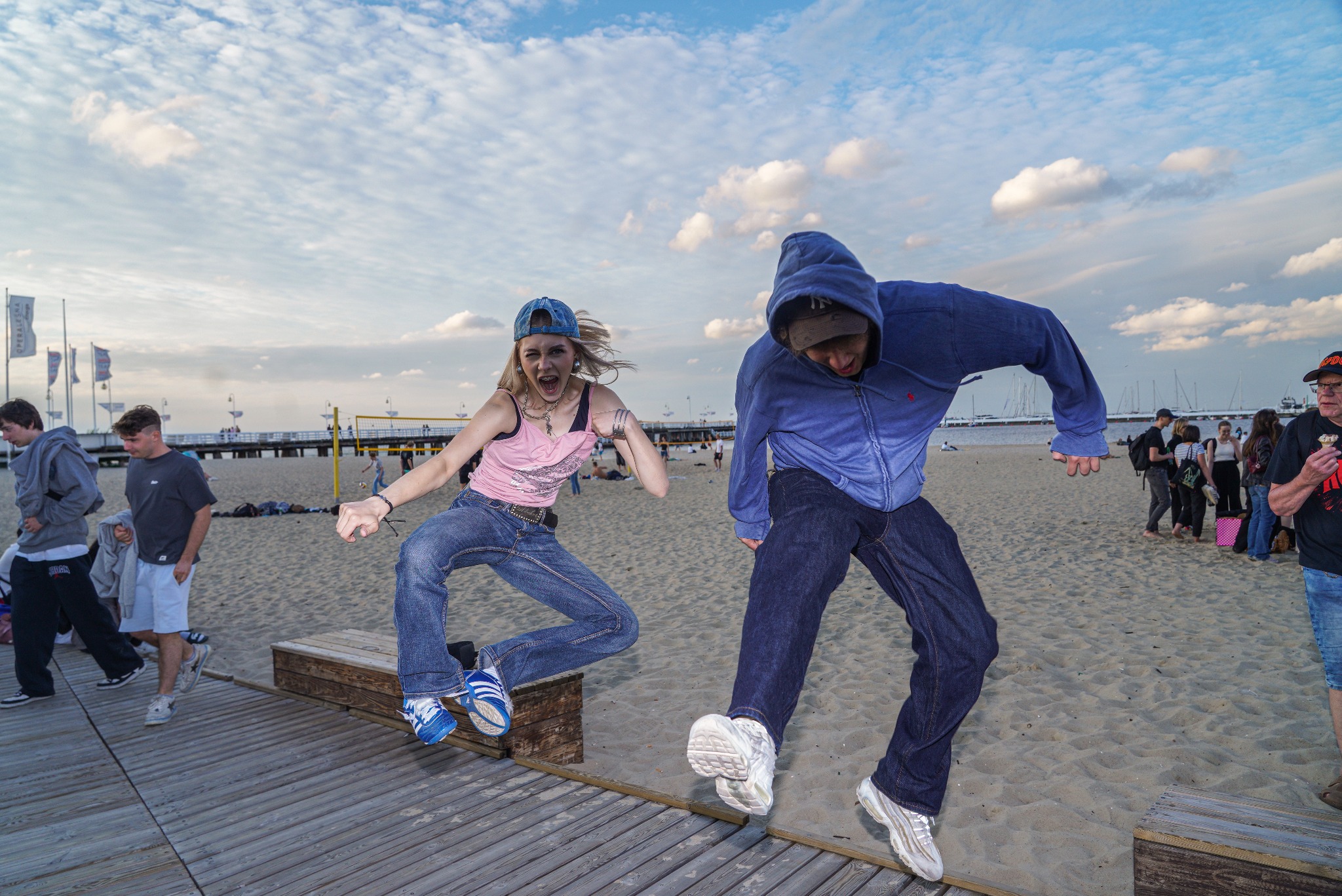 Dwie skaczące osoby na tle plaży i mola w słoneczny dzień. Dziewczyna w różowym topie i chłopak w bluzie z kapturem w dynamicznych pozach. W tle widoczni ludzie i elementy plażowej infrastruktury.