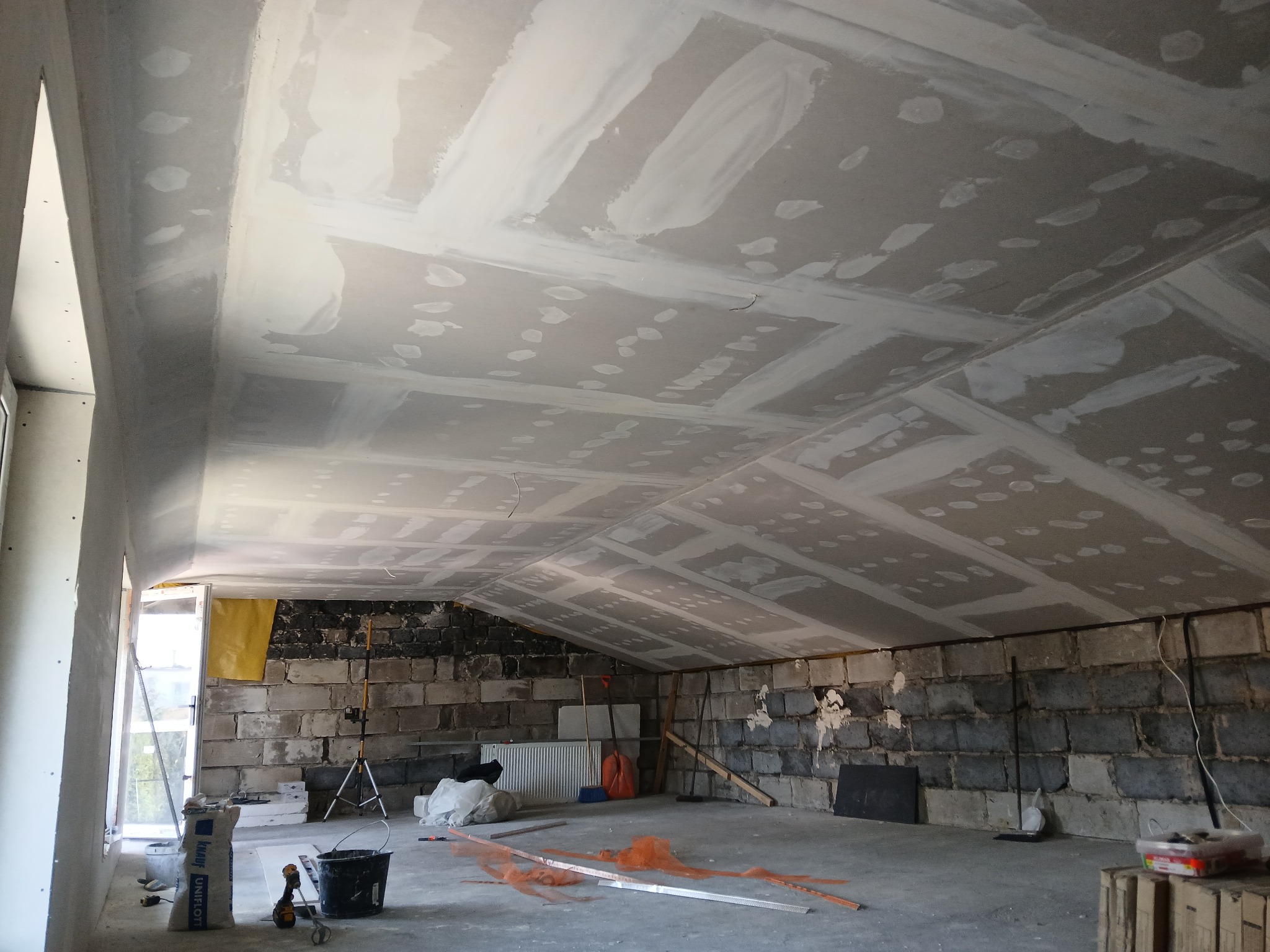 Poddasze w trakcie prac wykończeniowych: zamontowane płyty karton-gips na suficie, wstępnie wyszpachlowane. Widoczne narzędzia i materiały budowlane.