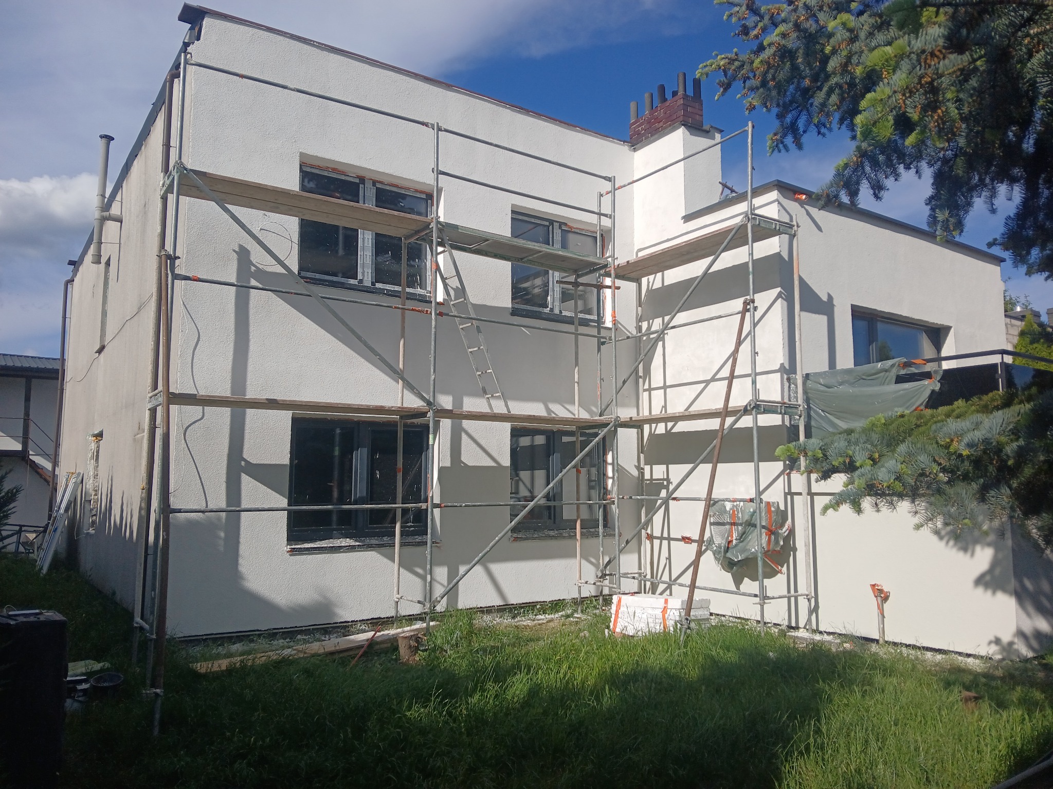 Biała fasada domu w trakcie odnawiania, z rusztowaniem i widocznym kominem. Świeżo pomalowana elewacja, błękitne niebo w tle, zieleń trawy na pierwszym planie.