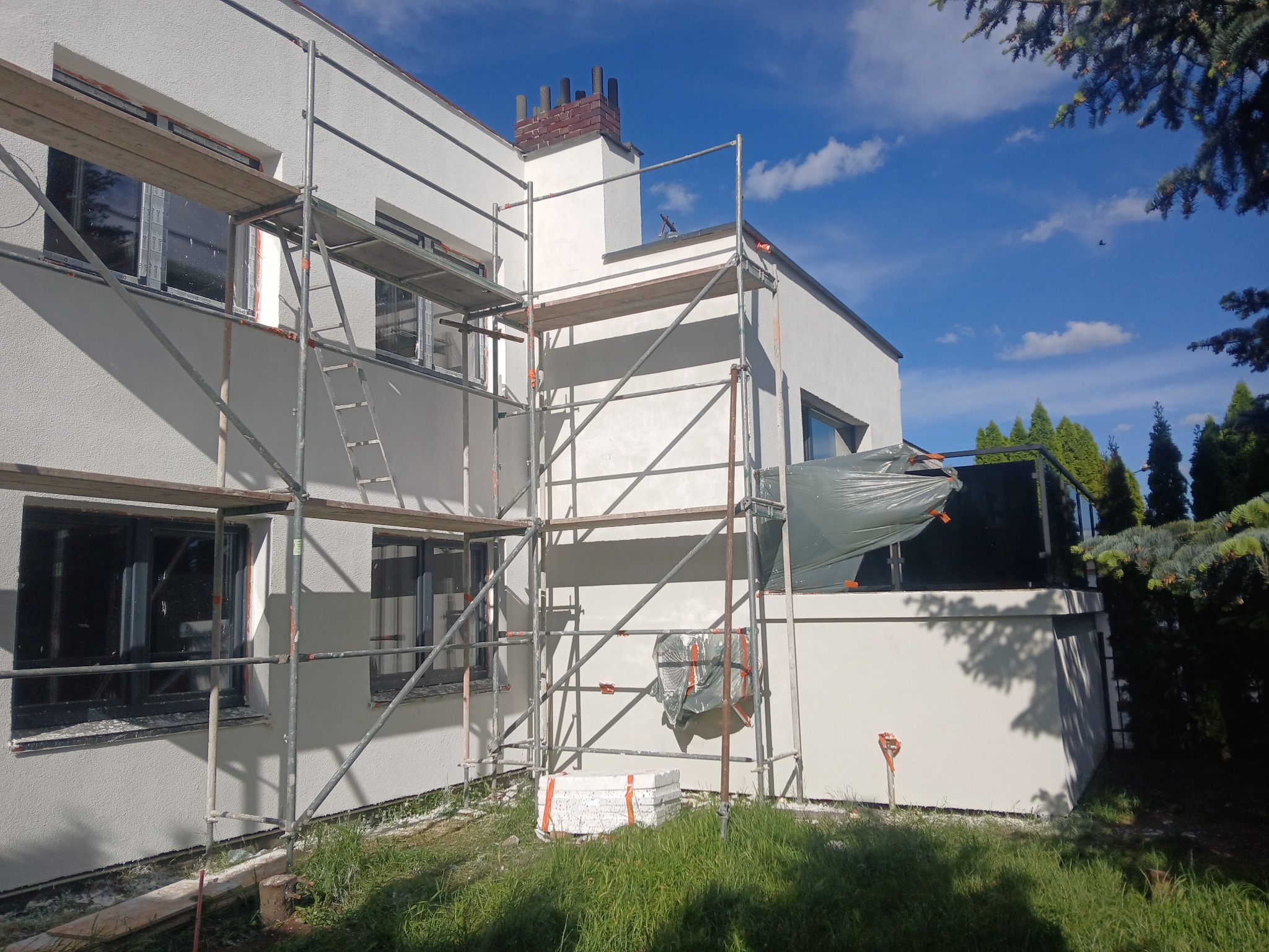 Biały dom z nową elewacją, w trakcie prac wykończeniowych. Widoczne rusztowania i materiały budowlane na trawniku. Słoneczny dzień, błękitne niebo.