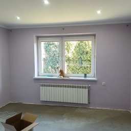 Le-Ma Bud Tomasz Gibas - Pokój po malowaniu na liliowo z kotem na parapecie. Widoczny grzejnik, osuszacz powietrza i karton. Świeżo pomalowane ściany, surowa podłoga.