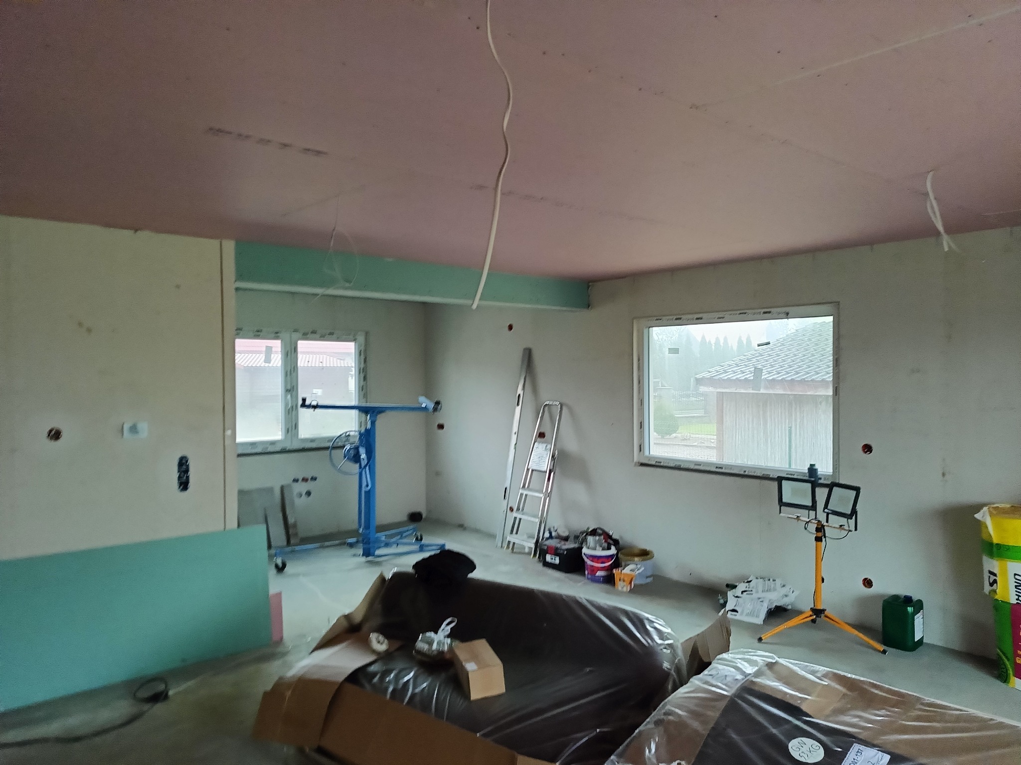 Wnętrze w trakcie montażu płyt gipsowo-kartonowych: widoczny podnośnik, płyty, drabina i oświetlenie budowlane. Sufit częściowo pokryty płytami w kolorze różowym.