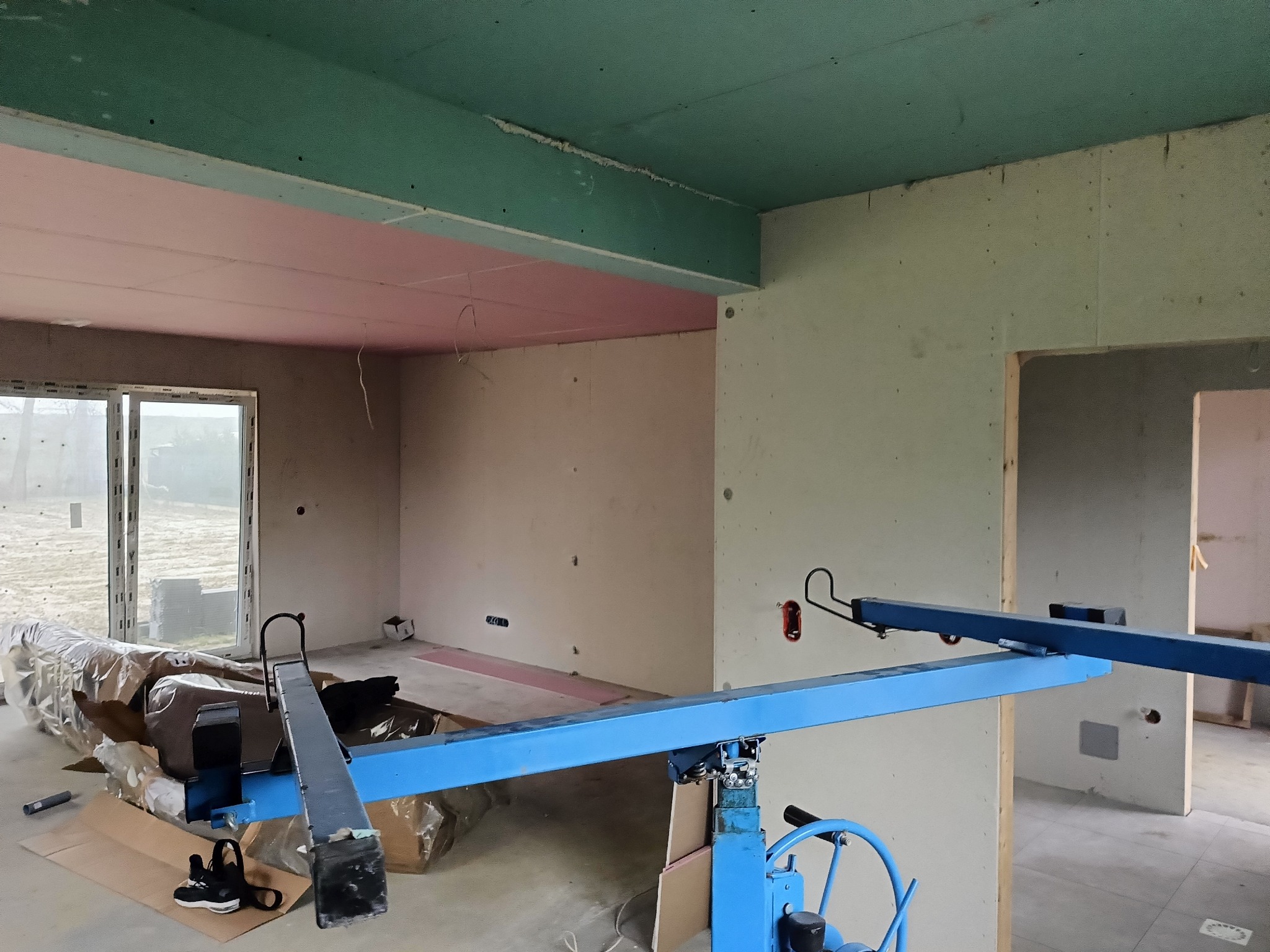 Wnętrze w trakcie montażu płyt gipsowo-kartonowych na suficie, z użyciem podnośnika. Widoczne surowe ściany i okno. Częściowo zakończone prace na suficie.