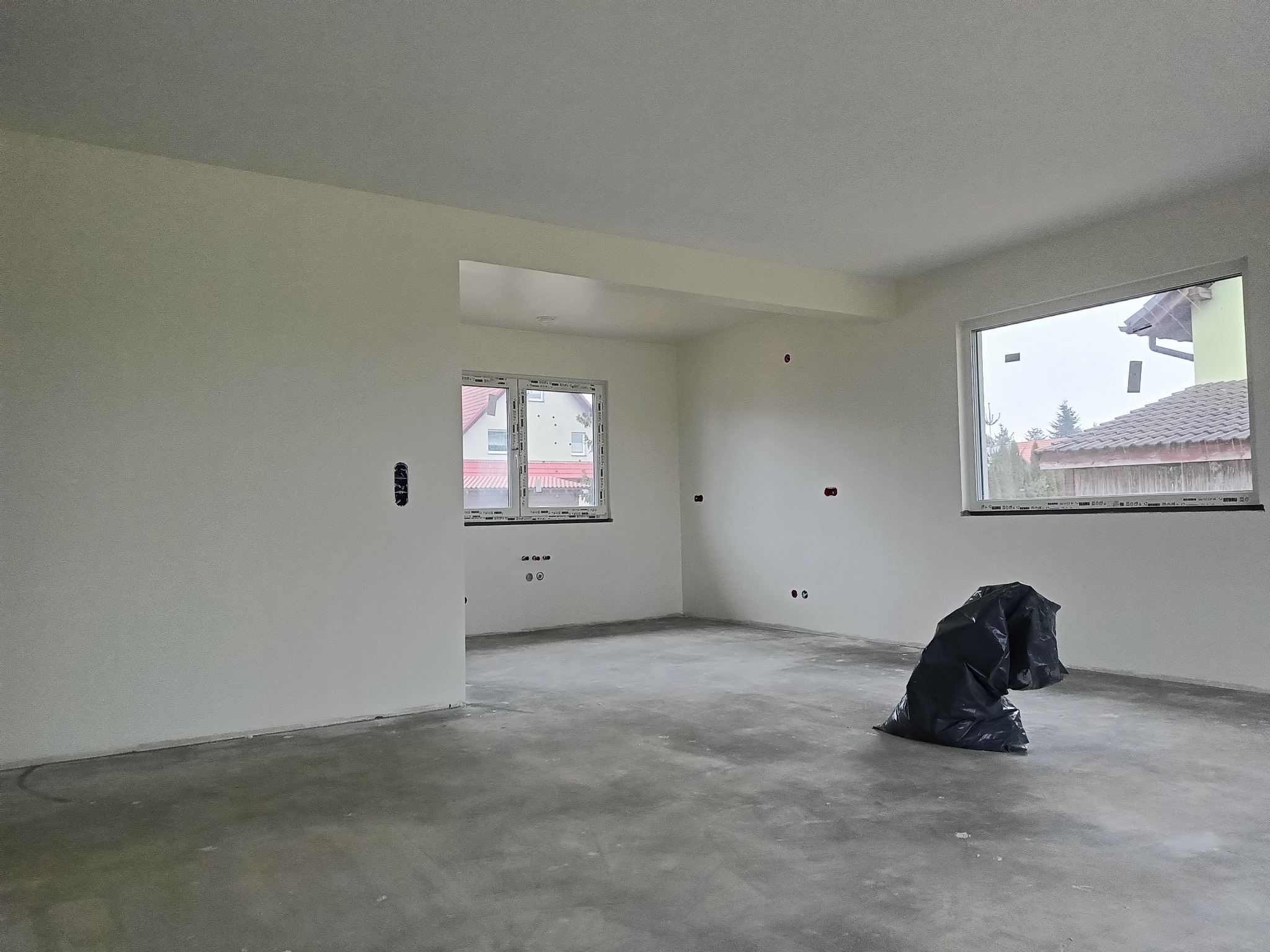 Wnętrze po malowaniu na biało, widoczne dwa okna, instalacja elektryczna i worek na śmieci. Ściany gładkie, surowe podłoże. Widok na domy przez okna.