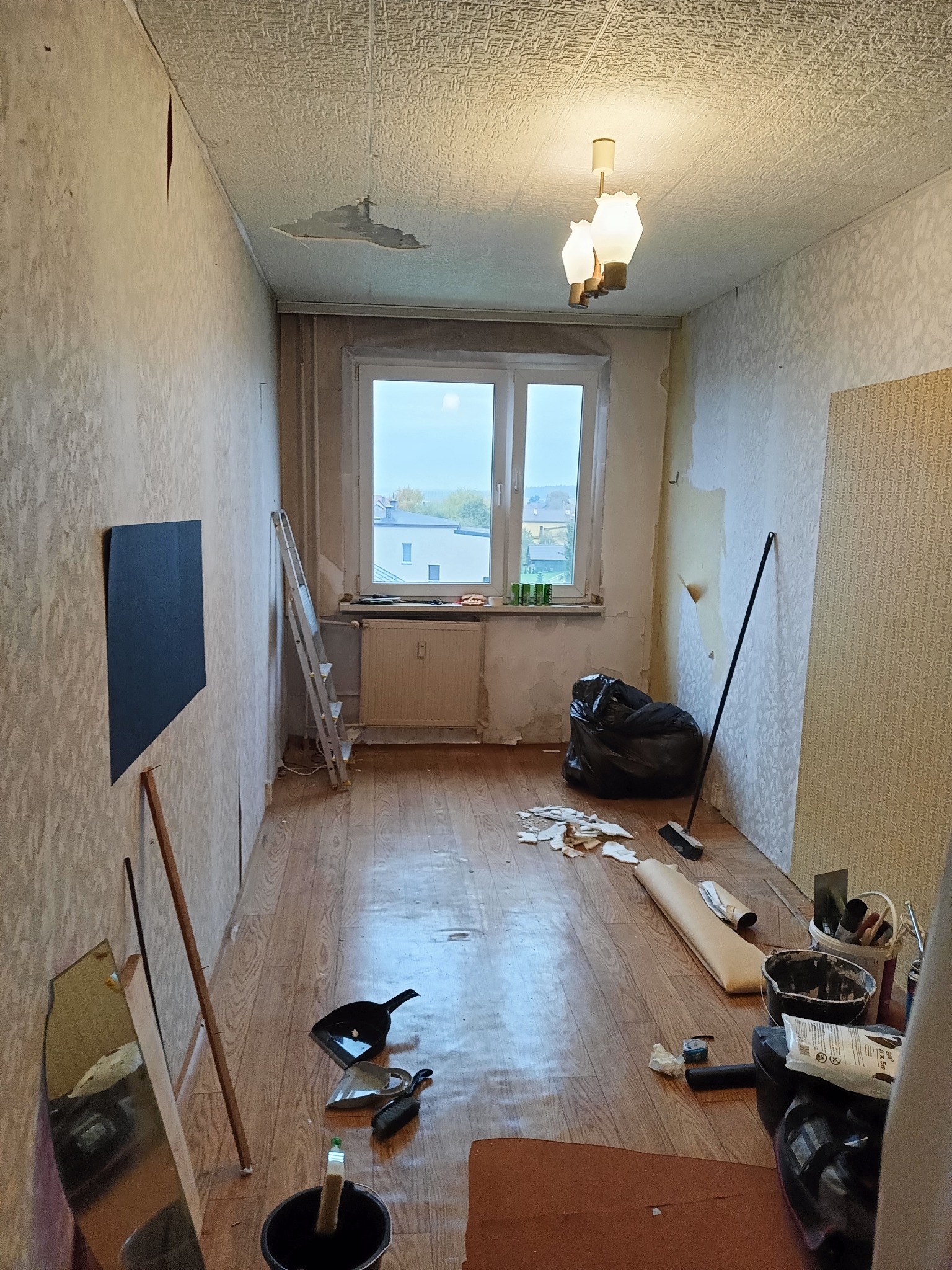 Pokój w trakcie remontu: zerwane tapety, widoczne narzędzia malarskie, drabina oparta o ścianę. Przy oknie czarny worek na śmieci. Podłoga częściowo zakryta folią.