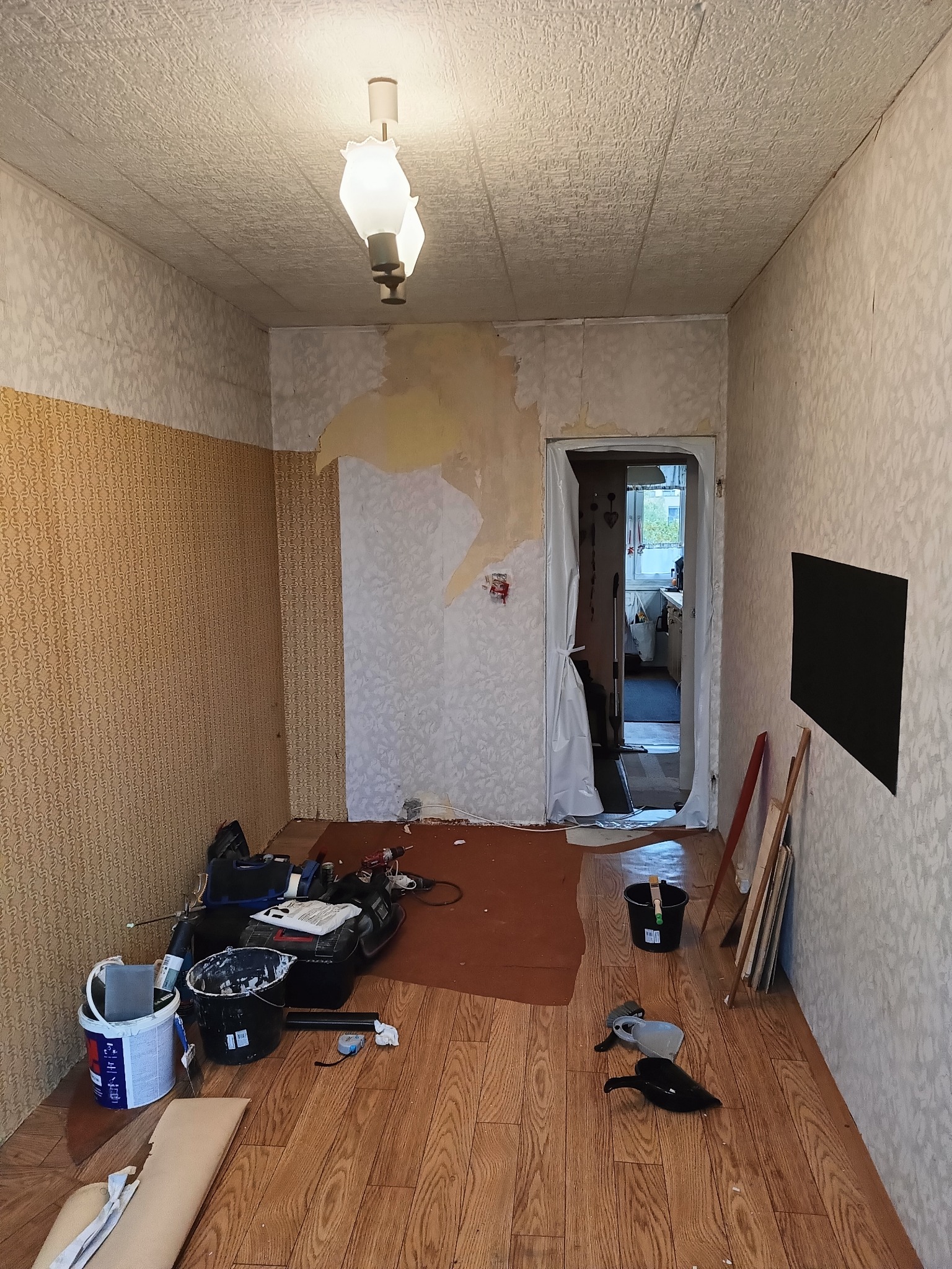 Pokój w trakcie remontu: zerwane tapety, przygotowane narzędzia malarskie, zabezpieczone drzwi folią. Widoczne ślady po starych tapetach i przygotowania do malowania.