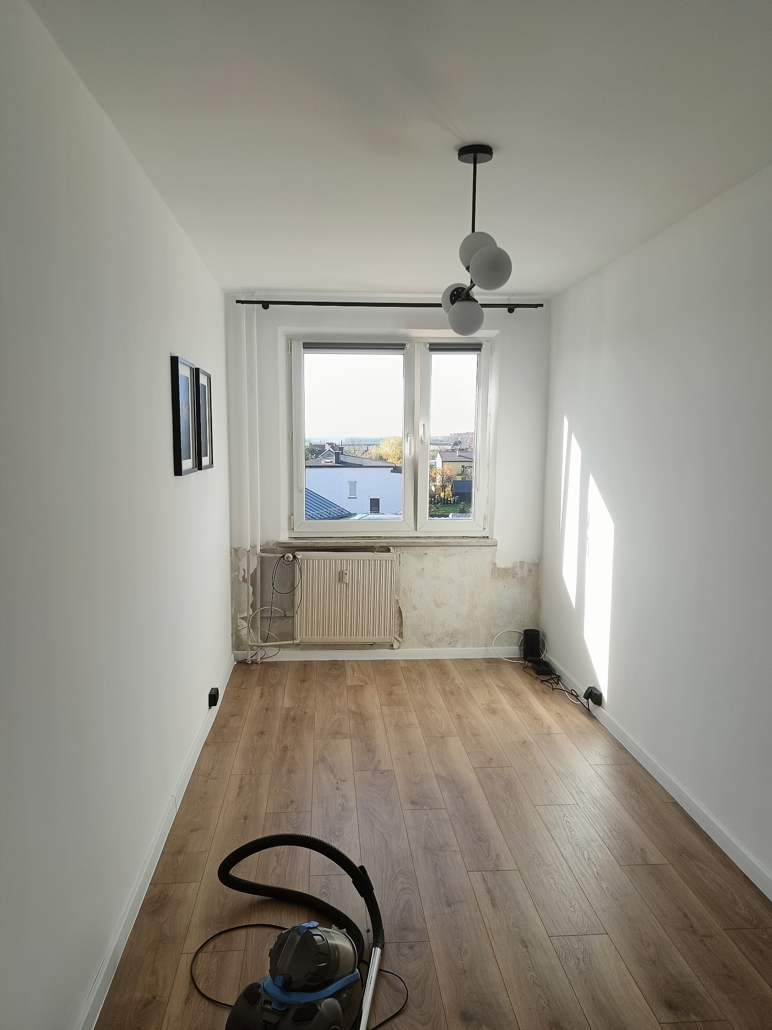 Pokój po szpachlowaniu, z widocznymi śladami prac przy oknie i grzejniku. Świeżo pomalowane ściany, drewniana podłoga i lampa sufitowa. Odkurzacz na pierwszym planie.