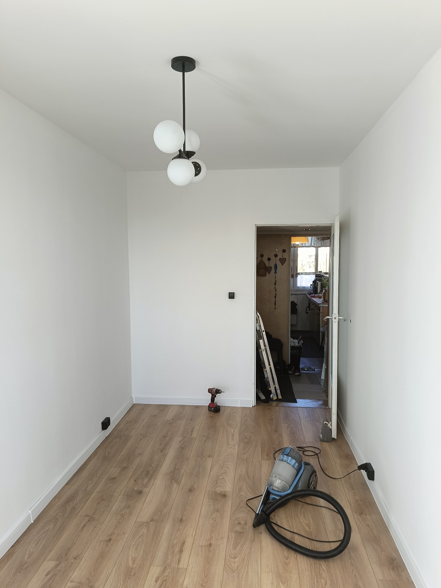 Jasne wnętrze po remoncie: białe ściany, drewniana podłoga, nowoczesna lampa. Widoczny odkurzacz i wiertarka, sugerujące prace wykończeniowe.