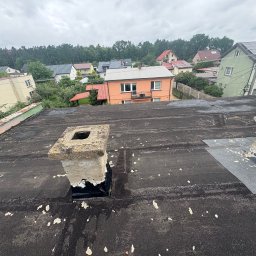 KamFix Kamil Borkowski - Czarny dach pokryty papą z widocznymi śladami napraw i odnowionym kominem na tle zabudowy jednorodzinnej w pochmurny dzień.