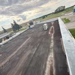 KamFix Kamil Borkowski - Praca na dachu płaskim, widoczny pracownik w oddali, sprzęt i klimatyzator. Dach pokryty papą, w tle stacja benzynowa i pochmurne niebo.