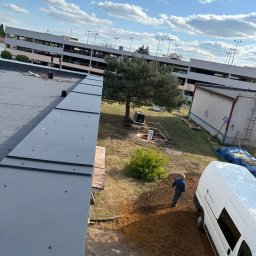 KamFix Kamil Borkowski - Szary dach z blachy na tle parkingu, drzew i nieba. Widoczny fragment dachu, fragment ziemi z pracownikiem i samochód dostawczy. Ujęcie z góry.