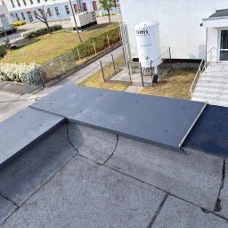 KamFix Kamil Borkowski - Szary, płaski dach pokryty papą z obróbką blacharską na krawędzi, budynek w tle. Widoczne ślady po naprawach.