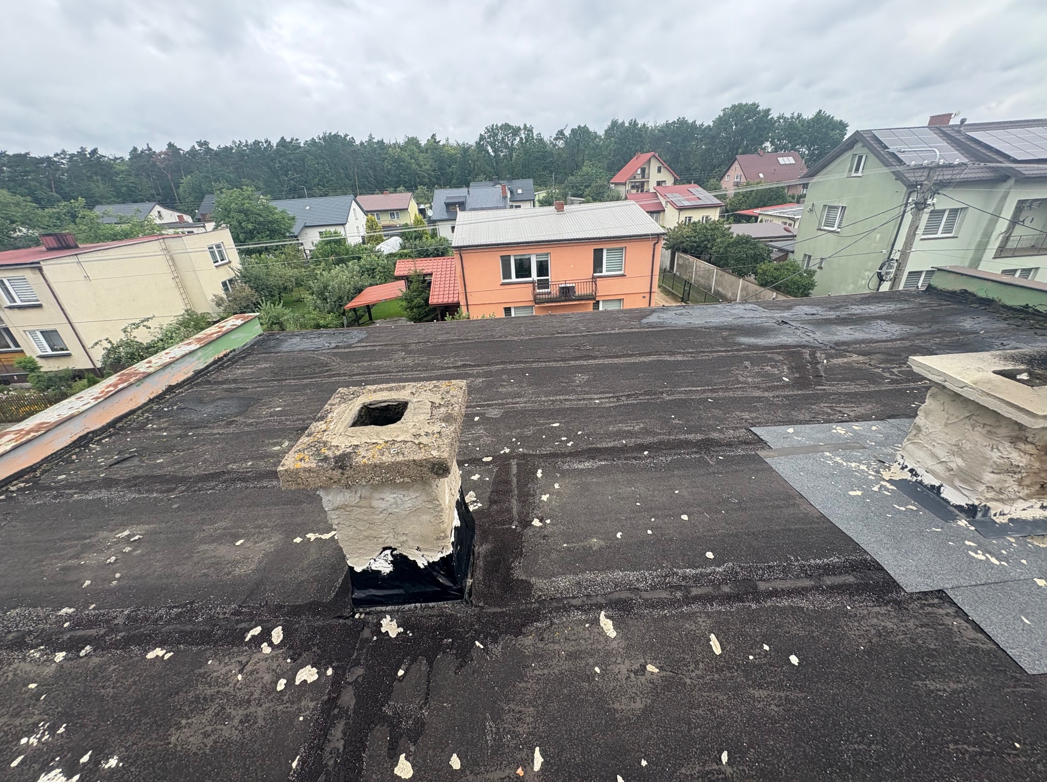 Czarny dach pokryty papą z widocznymi śladami napraw i odnowionym kominem na tle zabudowy jednorodzinnej w pochmurny dzień.