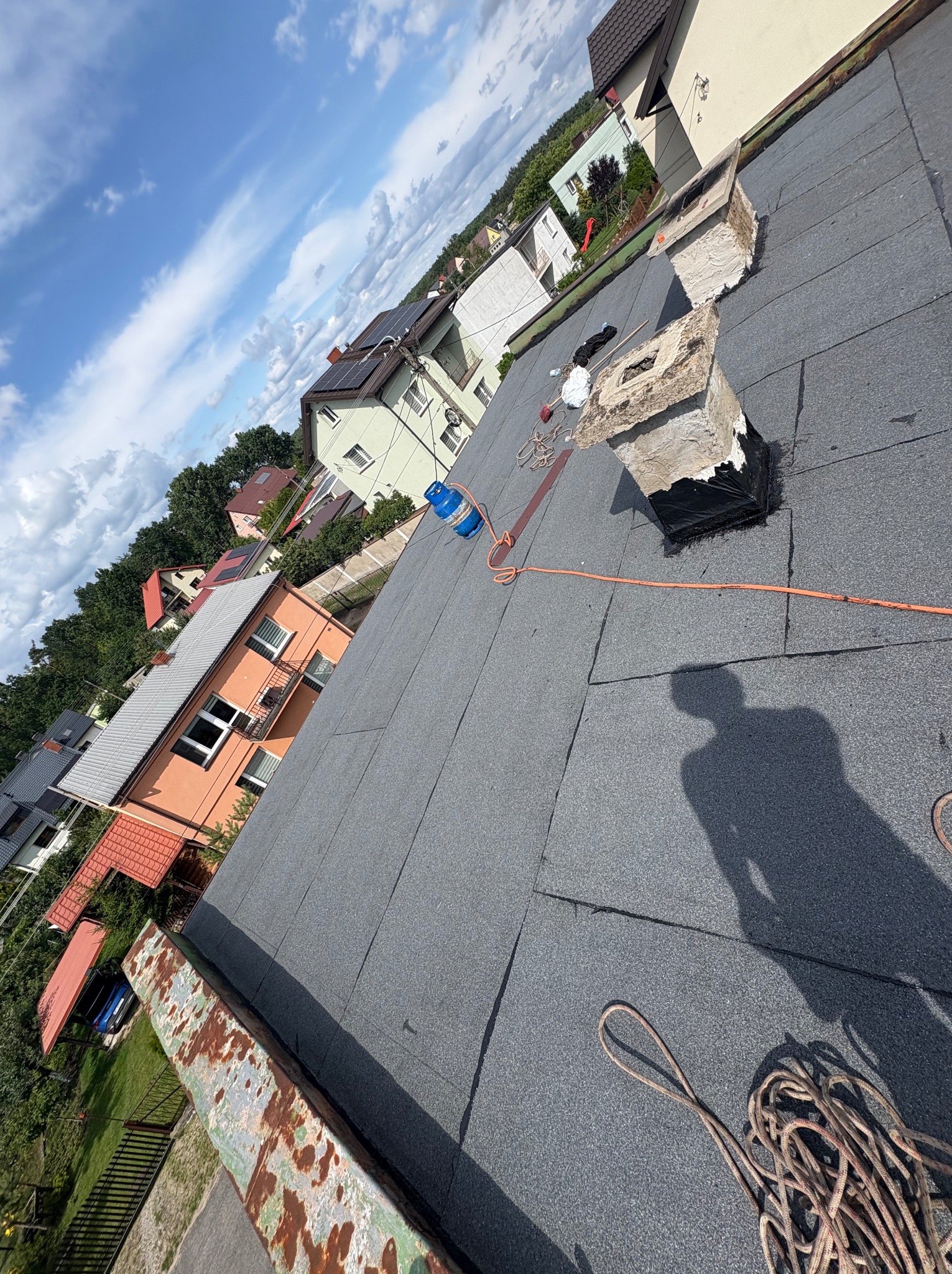 Szary, płaski dach pokryty papą termozgrzewalną, z widocznymi kominami i shadow postaci ludzkiej. W tle zabudowa jednorodzinna i błękitne niebo.