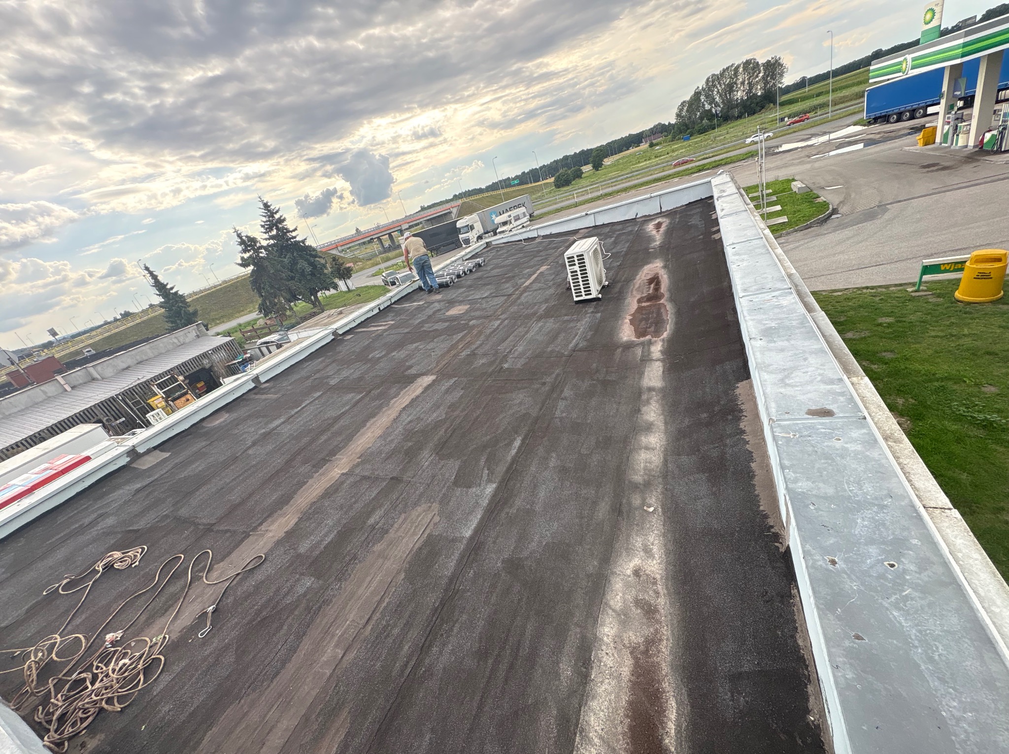 Praca na dachu płaskim, widoczny pracownik w oddali, sprzęt i klimatyzator. Dach pokryty papą, w tle stacja benzynowa i pochmurne niebo.