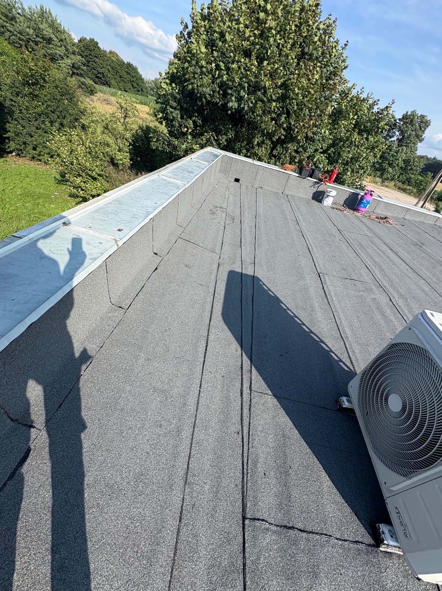 Szary dach pokryty papą termozgrzewalną z widocznym klimatyzatorem i narzędziami. Cień osoby robiącej zdjęcie. Tło: zieleń drzew i błękitne niebo.