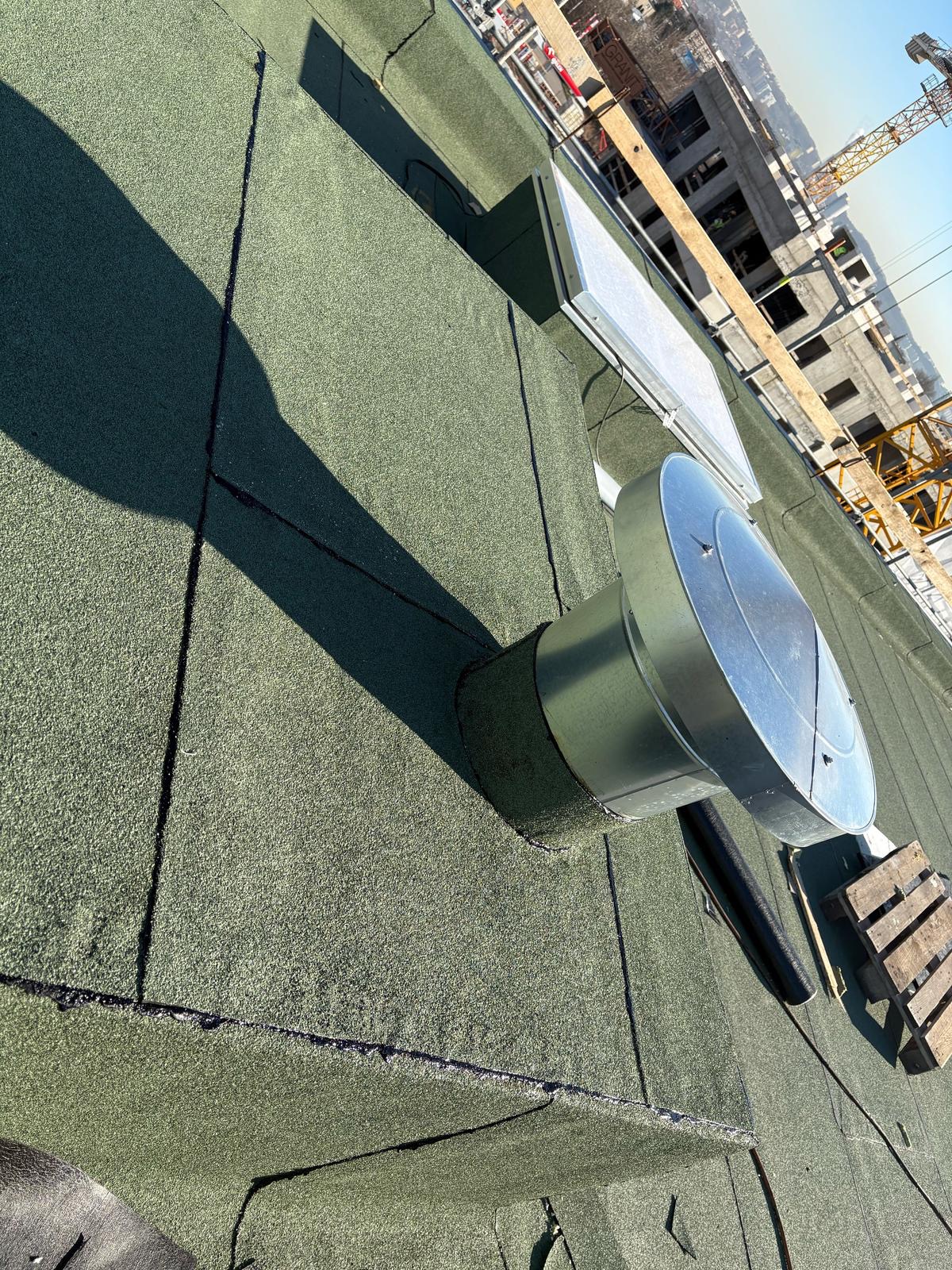 Zielony dach z papy termozgrzewalnej z wentylacją kominową, widok z góry, w tle budowa i dźwig. Paleta drewniana leży na dachu.