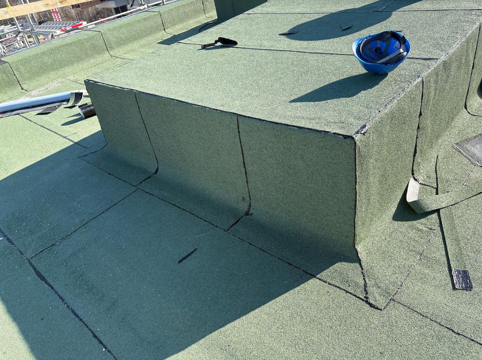 Zielony dach pokryty papą termozgrzewalną, widoczny niebieski kask i narzędzia dekarskie. Wykończony fragment dachu z detalami łączeń.