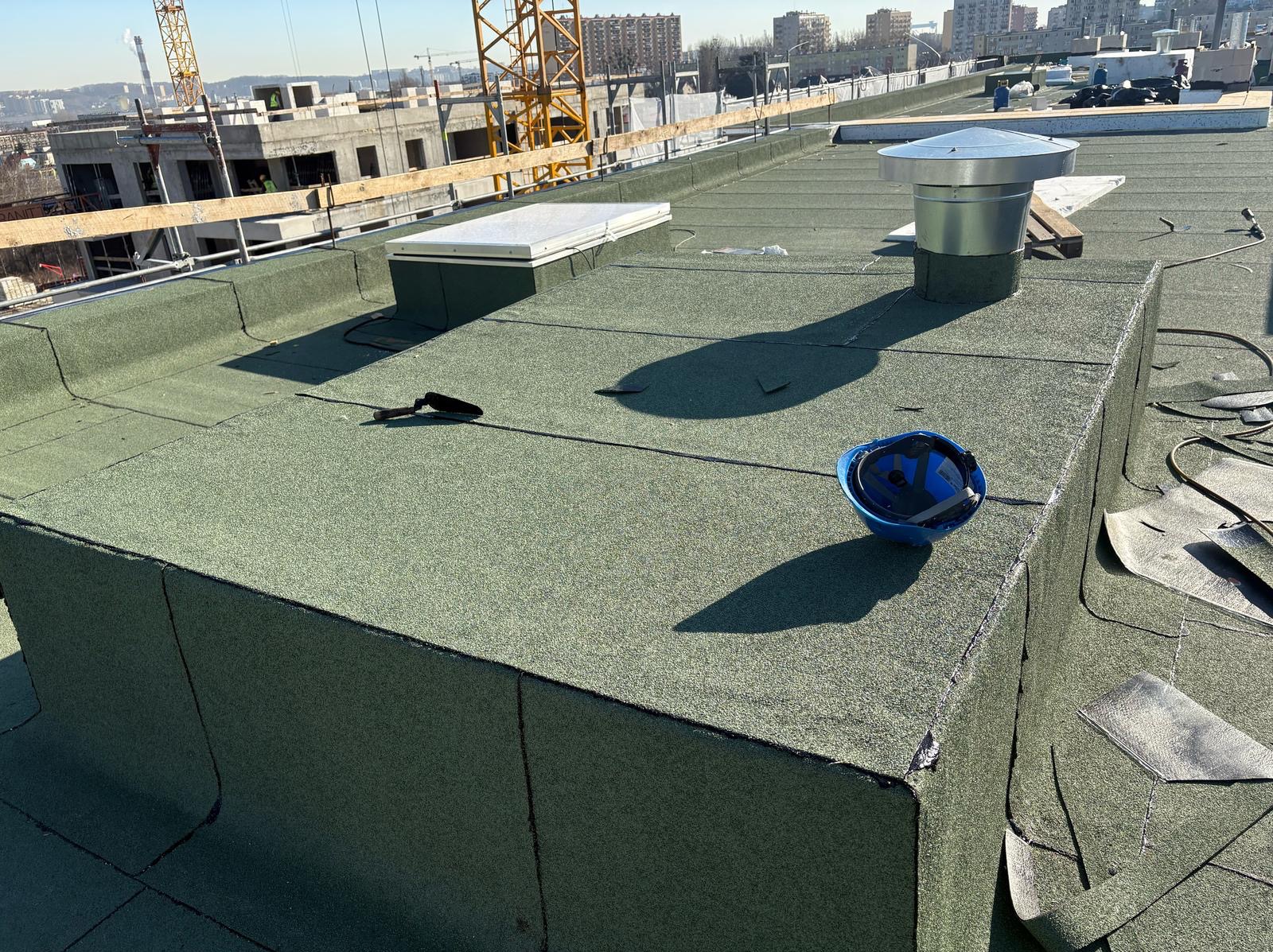 Zielony dach pokryty papą termozgrzewalną z kominem wentylacyjnym, włazem dachowym i kaskiem budowlanym. W tle widoczne budynki w trakcie budowy i żurawie.