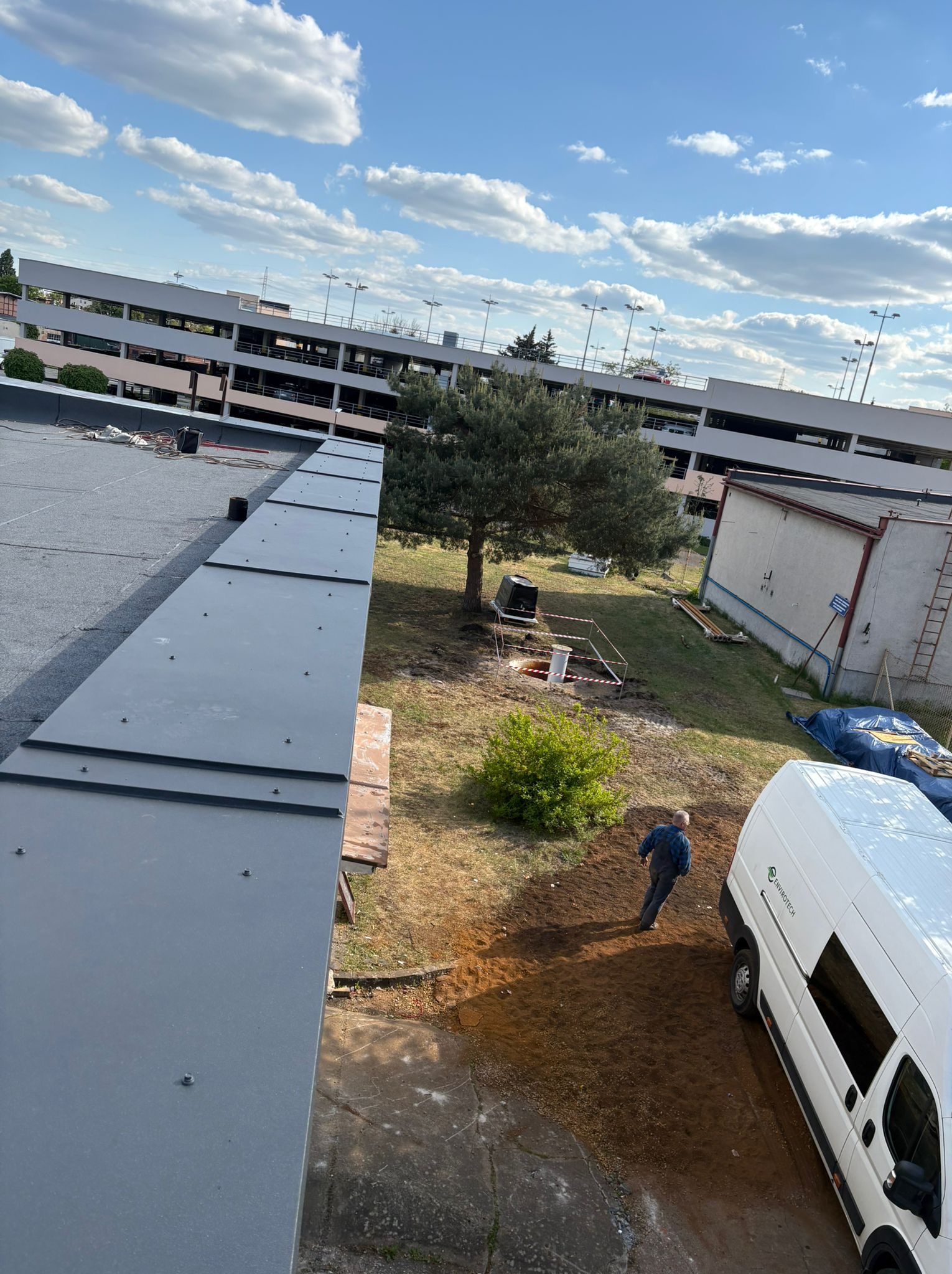 Szary dach z blachy na tle parkingu, drzew i nieba. Widoczny fragment dachu, fragment ziemi z pracownikiem i samochód dostawczy. Ujęcie z góry.