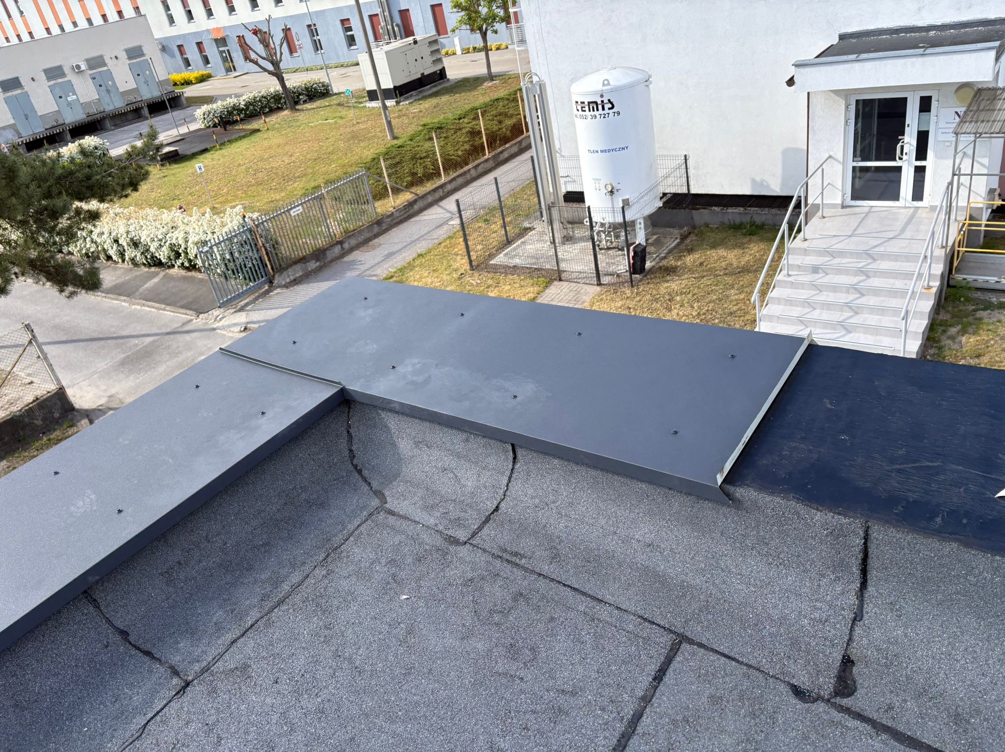 Szary, płaski dach pokryty papą z obróbką blacharską na krawędzi, budynek w tle. Widoczne ślady po naprawach.