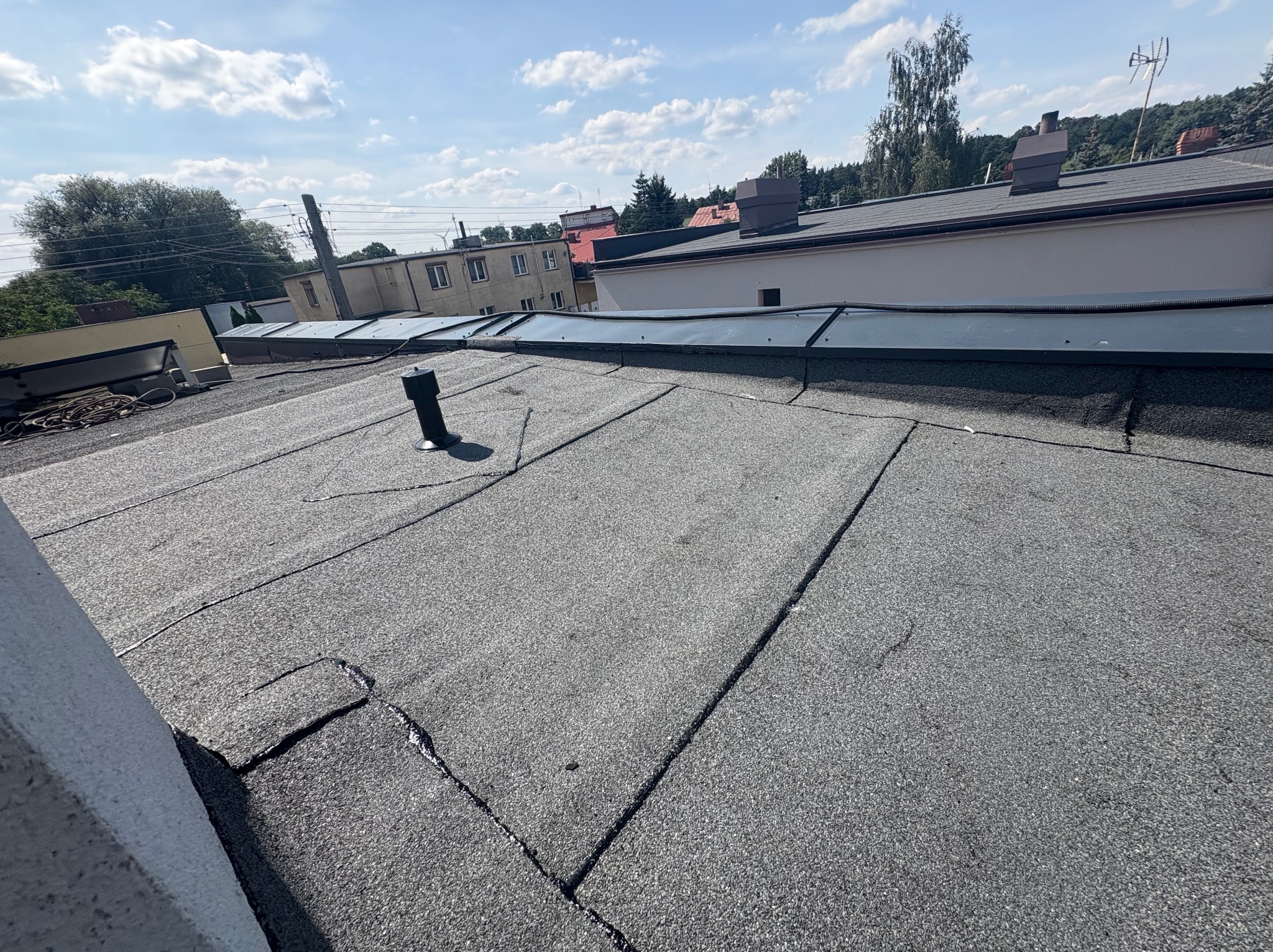 Szary, płaski dach pokryty papą, z kominkiem wentylacyjnym. Widoczne złączenia papy. W tle budynki i drzewa pod błękitnym niebem.