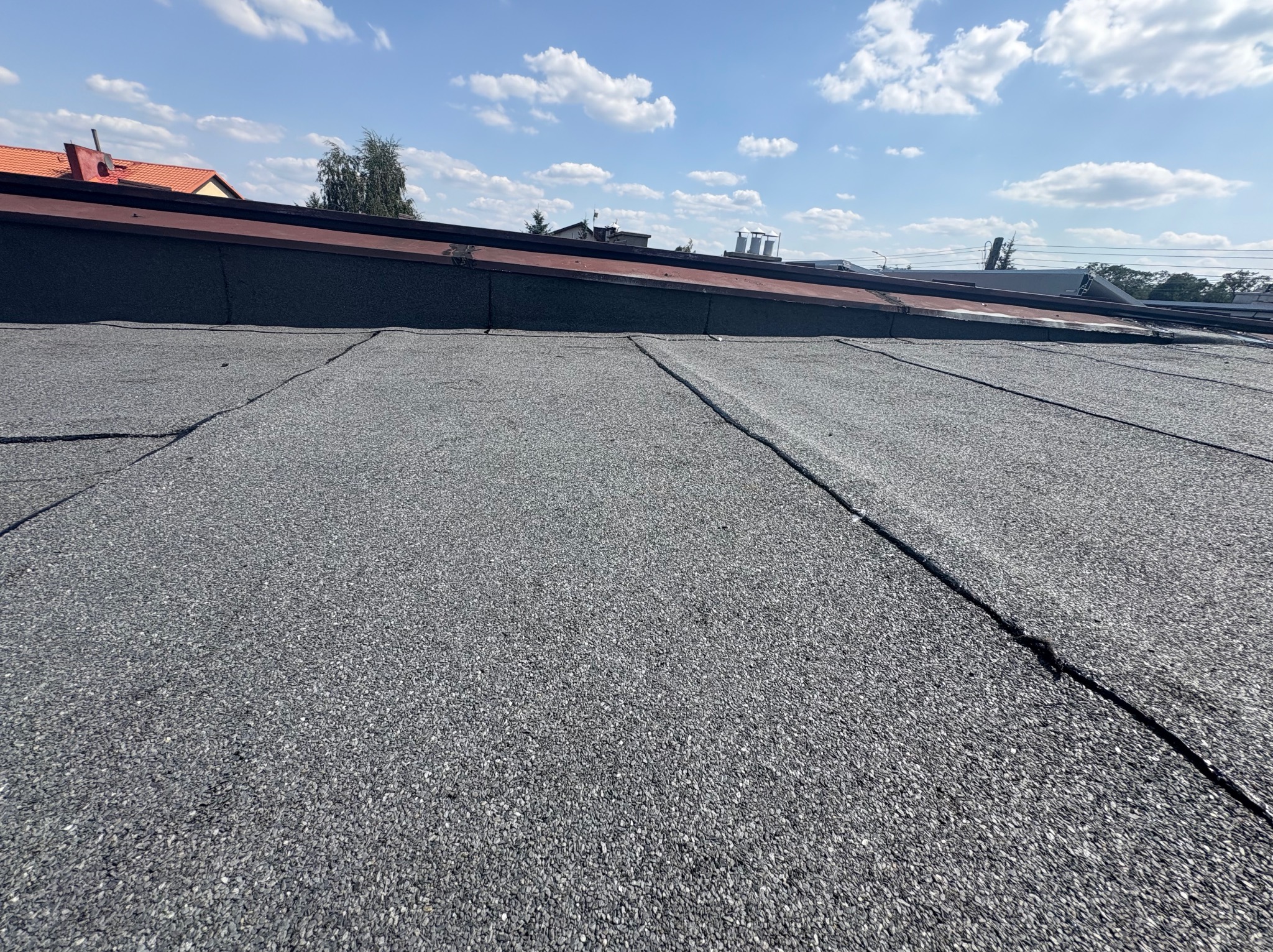 Szczegółowe ujęcie dachu pokrytego papą bitumiczną na tle błękitnego nieba z chmurami. Widoczne łączenia papy. W tle fragmenty budynków i zieleni.