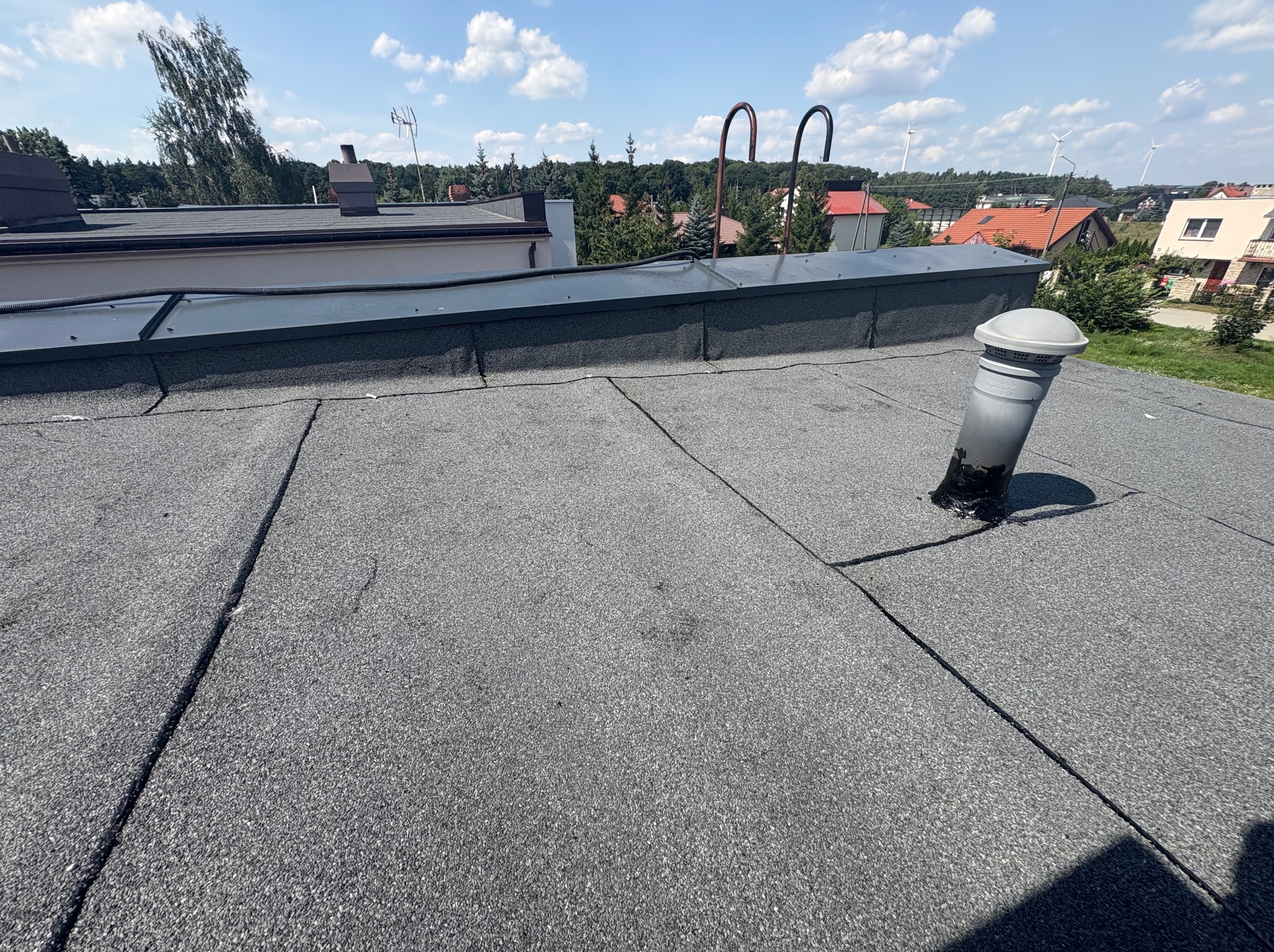 Szary, płaski dach pokryty papą z kominkiem wentylacyjnym. W tle budynki mieszkalne i turbiny wiatrowe. Widok z góry na pokrycie dachowe i otoczenie.