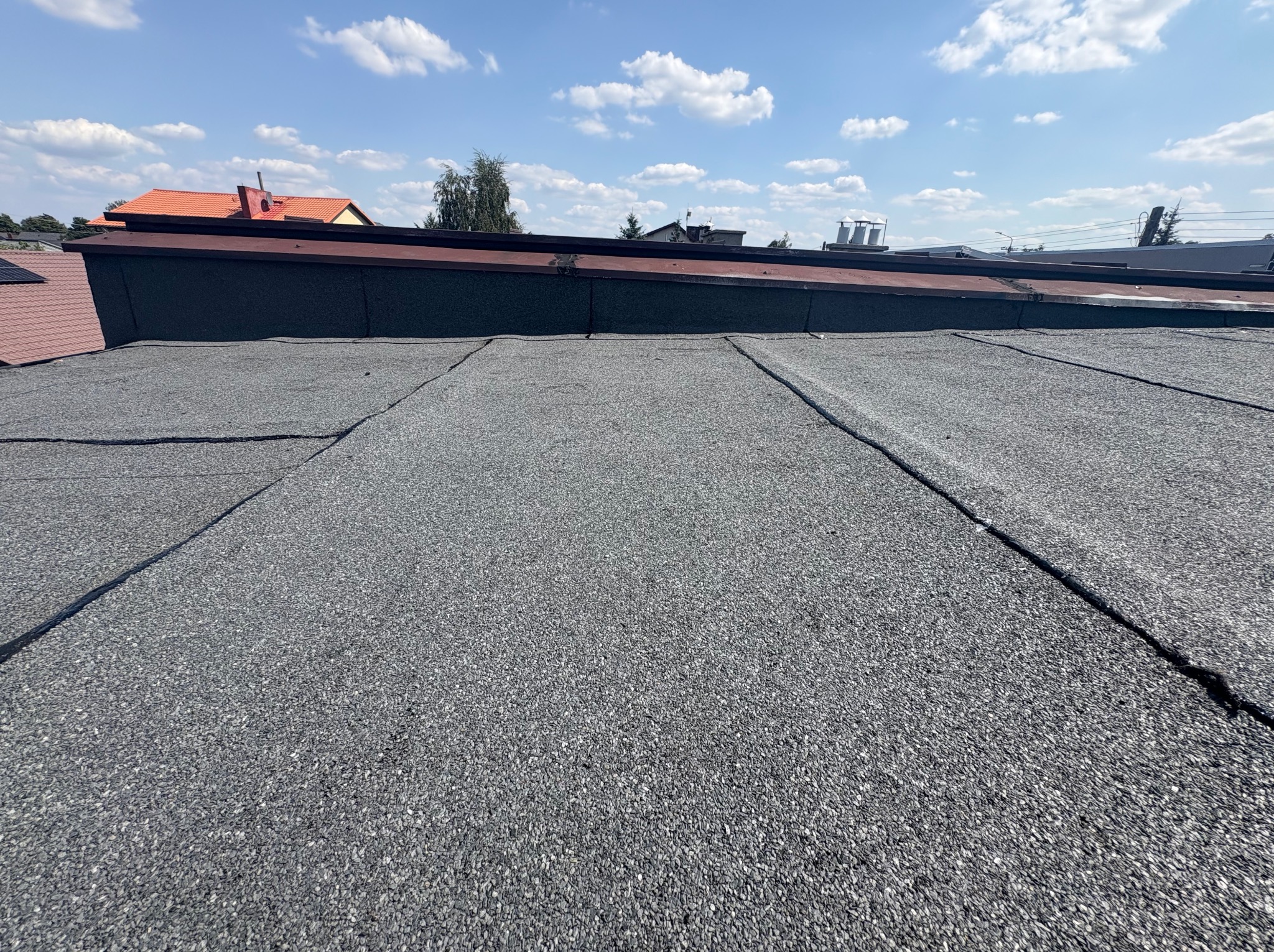 Szary dach pokryty papą bitumiczną w słoneczny dzień. Widoczne łączenia papy i fragmenty innych dachów w tle. Tekstura papy wyraźnie widoczna.