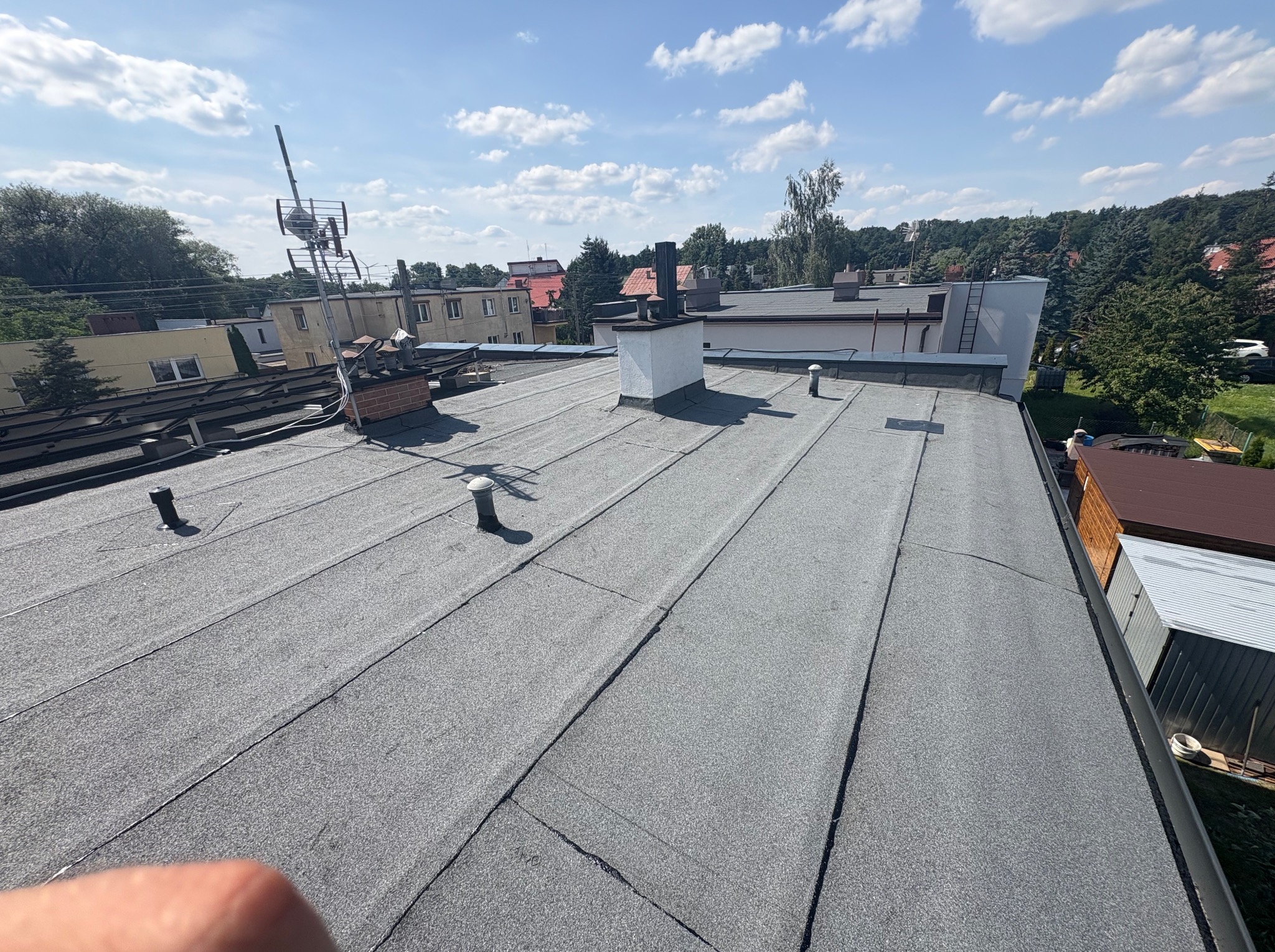 Dach pokryty papą termozgrzewalną z kominem i anteną na tle zabudowań i drzew. Widoczne łączenia papy. W tle błękitne niebo z chmurami.