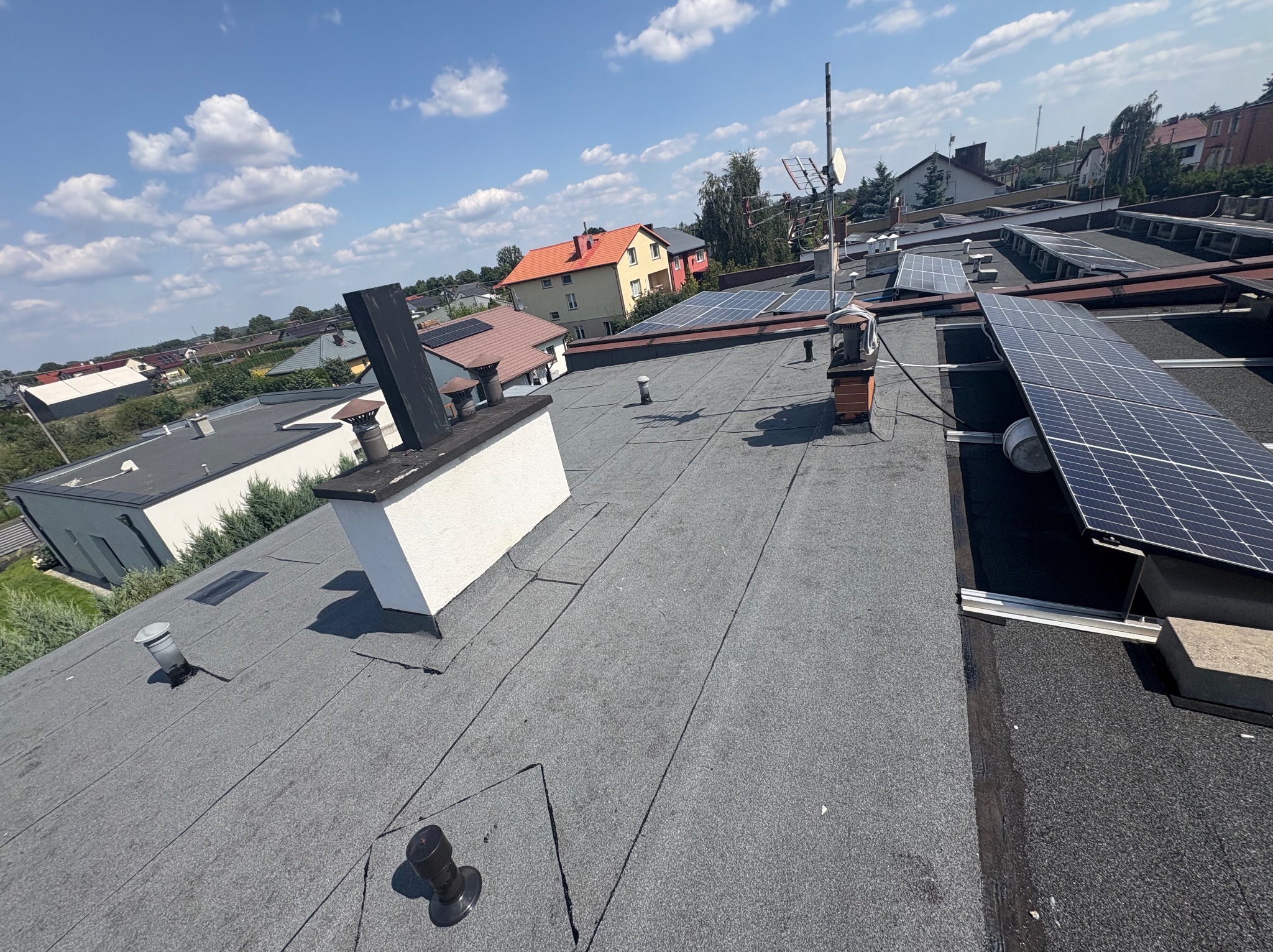 Dach pokryty papą termozgrzewalną z panelami fotowoltaicznymi i kominem wentylacyjnym na tle zabudowy miejskiej w słoneczny dzień.