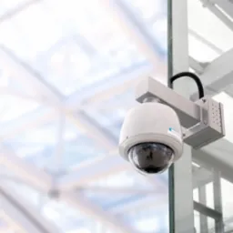 Kopułkowa kamera monitoringu CCTV zamontowana na szklanej ścianie w nowoczesnym budynku z metalową konstrukcją dachu w tle. Widoczny kabel zasilający.